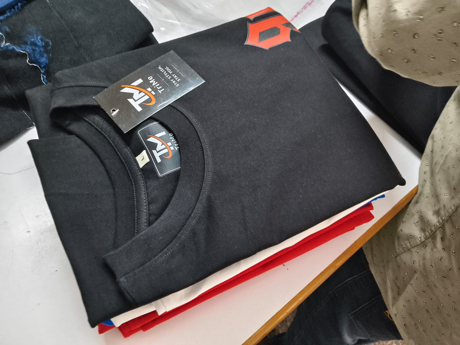 Folded black TRI/ME tee on a stack of tees